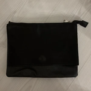 Svart kuvertväska  - Stilren svart kuvertväska som kan bli en axelrem väska. det är både ett skinnande material och mocka material. Har en stor huvudficka som går att dra igen med dragkedja och sedan en annan ficka som har en magnet som fäste. Den är från H&M  och är i bra skick, men dock har Moca materialet slitit lite då magnetkhnappen är under, se sista bild.