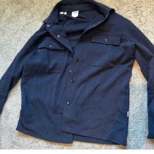 Mörkblå overshirt från Selected Homme - Snygg mörkblå overshirt från Selected Homme i bomull. Modellen har två bröstfickor med lock och tryckknappar, klassisk krage och långa ärmar. Perfekt att slänga över en t-shirt för en chill look.