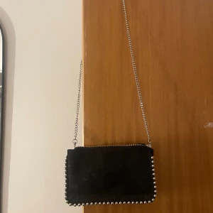 Svart väska  - Snygg svart axelväska i mocka med silverfärgad kedja och nitar längs kanterna. Väskan har dragkedja upptill och flera fack inuti. Använd ett fåtal gånger och är i fint skick!
