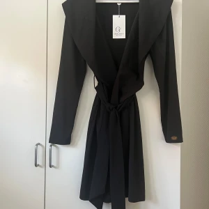 Svart kappa från Chiara Forthi - Stilren svart kappa från Chiara Forthi med bred krage och knytskärp i midjan. Långärmad modell med snyggt fall och minimalistisk design. Perfekt för dig som gillar en clean och elegant look.