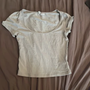 Ljusbeige croppad topp med kort ärm - Enkel och stilren ljusbeige topp med croppad passform och korta ärmar. Djup rundad u-ringning framtill ger en snygg och avslappnad vibe. Perfekt att matcha med jeans eller kjol för en clean look.