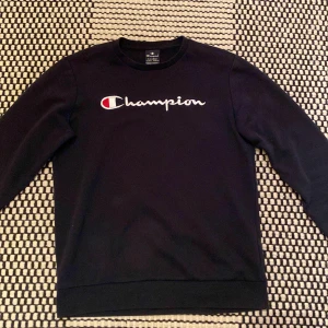 Mörkblå Champion sweatshirt cm 162/167 - Mörkblå sweatshirt från Champion i storlek X-Large, med broderad logga på bröstet och ärmen. Klassisk crewneck-modell med långa ärmar och ribbade muddar. Perfekt för en chill och sportig stil. Den är lite nopprig på vissa ställen (se bilder). Mjukt vadderad på insidan av hela tröjan. Hör gärna av er vid frågor!