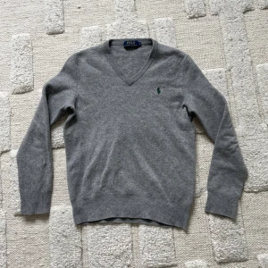 Grå v-ringad tröja Polo Ralph Lauren - Klassisk grå v-ringad tröja från Polo Ralph Lauren i mjuk ullmix. Tröjan har lång ärm, ribbade muddar och den ikoniska gröna broderade loggan på bröstet. Perfekt för lager-på-lager och enkel att matcha med jeans eller chinos.har krymt lite i tvätten