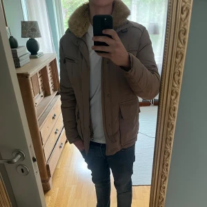 Beige dunjacka från Hetregó med fuskpäls - Säljer en beige dunjacka från Hetregó med mysig fuskpäls i kragen. Jackan har flera fickor med dragkedja och knappar, quiltad insida och en snygg detalj med metallogo på ärmen. Perfekt för kalla dagar och riktigt skön att bära.