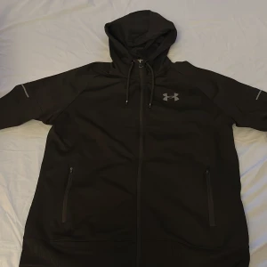Svart hoodie från Under Armour - Svart hoodie från Under Armour med huva och snörning. Hoodien har dragkedja framtill, två sidofickor med dragkedja och logga på bröstet samt text på ryggen. Tillverkad i polyester och har en loose passform, perfekt för träning eller chill. Jätte bra skick 9/10!🔥