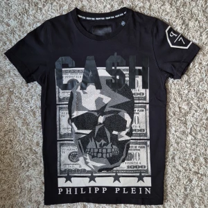 Svart t-shirt Cash Philipp Plein Nyskick - Svart t-shirt glittrande stor döskalle och dollartryck på framsidan samt texten 'CASH'. Märkeslogga på ärmen och nacken. Skön passform och cool design som sticker ut. Tillverkad i mjuk bomull. S/M