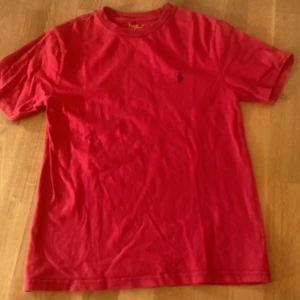  Polo Ralph Lauren röd tröja  - Röd t-shirt från Polo Ralph Lauren med klassisk blå broderad logga på bröstet. T-shirten har rund halsringning och korta ärmar. Tillverkad i mjuk bomull för en skön känsla och enkel stil.