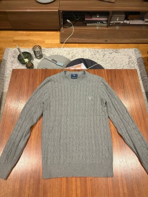 Grå kabelstickad tröja från Gant - Säljer en grå kabelstickad tröja från Gant i premium bomull. Tröjan har rund halsringning, ribbade muddar och diskret Gant-logo på bröstet. Perfekt för dig som gillar klassisk och stilren look.