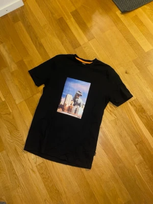 Svart BOSS t-shirt med tryck  - Svart t-shirt från BOSS med ett stort färgglatt tryck av en dinosaurie över stadssilhuett på bröstet. Klassisk rund halsringning med orange detalj på insidan av kragen. Skön och mjuk bomull som passar perfekt till jeans eller shorts.