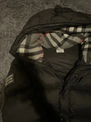 Svart pufferjacka från Burberry - Säljer en svart pufferjacka från Burberry med klassiskt rutigt foder i huvan. Jackan har dragkedja, tryckknappar och en tydlig Burberry London England-patch på ärmen. Perfekt för kalla dagar och riktigt snygg med sin oversized look.