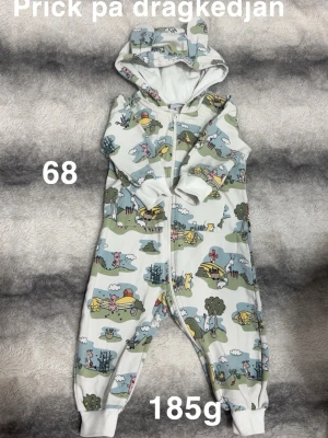 Mönstrad jumpsuit med dragkedja - Söt jumpsuit med huva och dragkedja framtill. Plagget har ett lekfullt mönster med djur och natur i blå, grön och vit färg. Muddar vid ärm- och benslut för extra komfort. Perfekt för mysiga dagar hemma.🌸 Använd två gånger. Liten prick på dragkedjan och lite missfärgat vid halsen. Storlek 68 