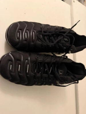 Nike Vapormax svarta sneakers - Säljer ett par svarta Nike Air Vapormax sneakers med cool, futuristisk design och genomskinlig sula. Skorna har snörning och detaljer i svart plast längs sidorna. Märkt med 'Vm Air' på hälen och Nike-logga på plösen.