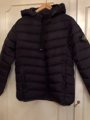 Svart pufferjacka från Stone Island - Säljer en svart pufferjacka från Stone Island med ikonisk patch på ärmen och huva. Jackan har quiltad design, dragkedja framtill och två sidofickor. Storlek M, passar M.