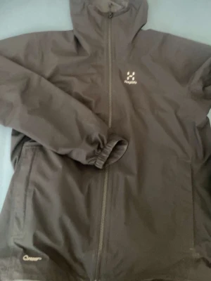 Svart vindjacka från Haglöfs - Säljer en svart vindjacka från Haglöfs i storlek M. Jackan har huva, hel dragkedja framtill och elastiska muddar vid ärmsluten. Tillverkad i lätt och slitstark polyester, perfekt för blåsiga dagar. Diskret Haglöfs-logga på bröstet och enkel, clean design.