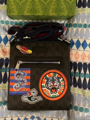 axelväska med patches - Säljer en svart axelväska med patches framtill, bland annat en tiger och planet. Väskan har justerbar axelrem i rött och blått, dragkedja framtill och detaljer i skinn. Perfekt för dig som vill sticka ut med en unik designerbag.