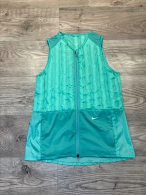 Nike Therma-FIT ADV - Snygg turkos dunväst från Nike Therma-FIT ADV med quiltad framsida och hel dragkedja. Västen har två stora fickor framtill och en liten Nike-logga på bröstet. Materialet är lätt och syntetiskt, perfekt för träning utomhus under kyliga dagar. Storlek M, passar S