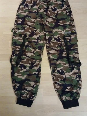 Camo cargopants med resår och detaljer. Från Temu! - Säljer ett par cargopants i klassiskt camouflage-mönster med gröna, bruna och svarta toner. Byxorna har resår i midjan och vid bensluten, stora fickor på sidorna och coola svarta banddetaljer. Perfekta för dig som gillar streetwear och vill sticka ut. Från Temu!