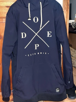 Mörkblå hoodie från Dope M - Snygg svart skid jacka från Dope med vit tryckt logga på bröstet och vita dragsnören i huvan. Utrustad med stor magficka och dragkedjor i sidorna för extra ventilation. Insidan är fodrad med mjukt material för skön komfort. Perfekt för chill eller utomhusaktiviteter.