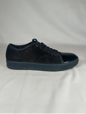 Svarta lanvins i mocka och lack - Snygga svarta lanvins med ovandel i mjuk mocka och glansig lackdetalj på tån. Skorna har snörning med svarta skosnören och metallöljetter. Platt sula och rund tå ger en clean och modern look. Perfekta till jeans eller kjol för en stilren vibe.