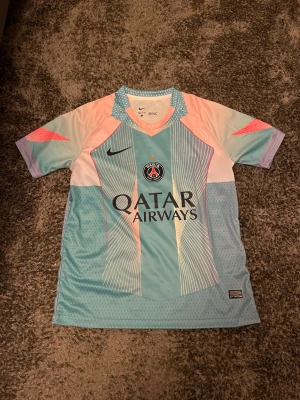 PSG x Nike pastell fotbollströja - Säljer en Paris Saint-Germain fotbollströja från Nike i snygga pastellfärger. Tröjan har ett unikt mönster i ljusblått, rosa och vitt med PSG-logga och Qatar Airways-tryck på bröstet. Tillverkad i lätt polyester med Dri-FIT-teknologi för bästa komfort på planen. 