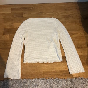 Vit långärmad topp med vågig kant - Säljer en vit långärmad topp med vågig kant nertill och ett diskret strukturerat mönster i tyget. Toppen har en rak passform och är tillverkad i ett mjukt material som känns skönt mot huden. Perfekt att styla med jeans eller kjol.