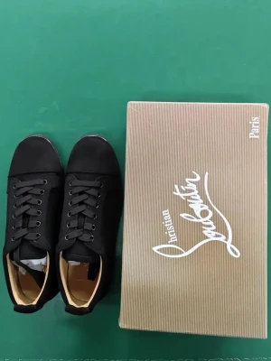 Svarta Christian Louboutin sneakers - Snygga svarta sneakers från Christian Louboutin med klassisk röd sula och diskret broderad logga på hälen. Skorna har snörning, rund tå och är tillverkade i en mix av skinn och syntet. Perfekta för dig som vill ha en stilren och lyxig look.