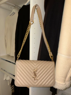 Beige quiltad axelväska från YSL AA - Säljer en beige quiltad axelväska från YSL med guldfärgad kedjerem och ikonisk YSL-logga framtill. Väskan är tillverkad i skinn och har ett elegant, stilrent utseende med v-format mönster på framsidan. Perfekt för dig som vill ha en lyxig och trendig accessoar.