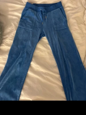 Blå velourbyxor Juicy Couture XS - Säljer ett par blå mjukisbyxor från Juicy Couture i velour. Byxorna har snörning i midjan, stora fickor fram och bak med knappdetaljer samt broderad JC-logga på ena fickan. Perfekta för en chill och trendig look.