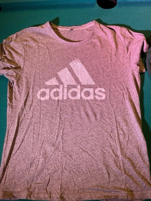 Grå Adidas t-shirt i XL - Snygg grå t-shirt från Adidas i storlek XL. T-shirten har klassisk rund hals och stort Adidas-logga framtill. Tillverkad i mjukt material som känns skönt mot huden. Perfekt för dig som gillar sportig och avslappnad stil.