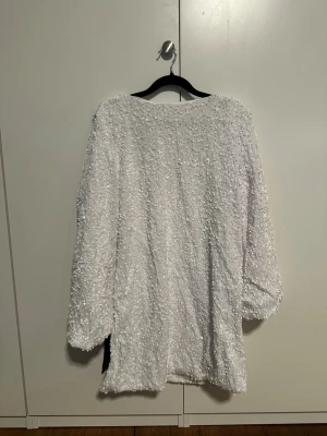 Vit paljettklänning med svart detalj - En vit kortklänning täckt av glittriga paljetter och med en djup v-ringning på ryggen. Klänningen har långa ärmar och en bred svart satinliknande band du kan knyta till en rosett på ryggen. Har aldrig används.