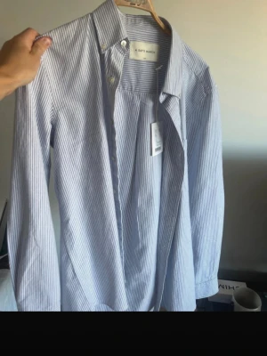 Randig skjorta från A Day's March - Snygg skjorta från A Day's March i vitt och blått med tunna vertikala ränder. Klassisk krage med knappar och lång ärm. Skjortan är tillverkad i bomull och har en clean, tidlös look som funkar till många olika stilar.