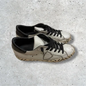 Philippe Model skor - Vita Philippe Model skor med svarta detaljer på sulan. Skorna kommer i stl 40 och är i bra skick, de mesta slitningarna är design. Undersulan är dock lagad med lim eftersom den brast lite vid hälen, kom privat för bild. Hör av er vid frågor!