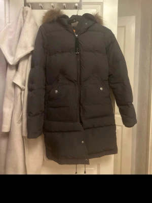 Svart lång dunjacka Parajumpers - Lång svart dunjacka från Parajumpers med stor huva och pälsdetalj. Jackan har två stora fickor framtill, dragkedja och tryckknappar. Klassisk patch på ärmen och justerbar nederkant med snörning. Perfekt för kalla dagar. Använd 2-3 gånger. Pris kan diskuteras!