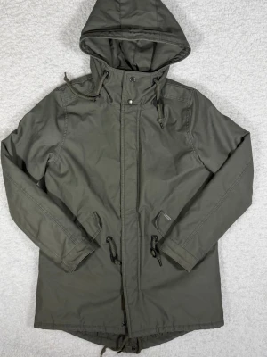 Olivgrön parkajacka från Carhartt - Säljer en olivgrön parkajacka från Carhartt med huva och dragsko i midjan. Jackan har quiltat foder, två stora fickor framtill och stängs med dragkedja och tryckknappar. Perfekt för dig som vill ha en stilren och funktionell jacka.