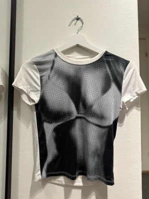 Vit t-shirt med svart tryck - Cool vit t-shirt med korta ärmar och ett svartvit grafiskt tryck framtill som ser ut som en muskulös överkropp. T-shirten har en klassisk rund halsringning och är gjord i ett mjukt material som känns skönt mot huden. Perfekt för dig som vill sticka ut med en unik look.