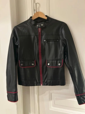 Svart skinnjacka/bikerjacket från Sisley - Svart skinnjacka från Sisley i storlek 44. Jackan har CADIG dragkedja framtill med röda detaljer, två fickor med tryckknappar och en bröstficka med dragkedja. Snygga röda sömmar vid ärmslut och fickor ger extra edge. Köpte i Rom, aldrig använt. Rekommenderar för personer som har XS eller S, sitter lite tajt på mig som brukar ha M