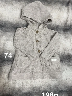 Beige stickad kofta med huva Newbie - Superfin beige stickad kofta från Newbie med huva och snygga flätade detaljer. Tre träknappar framtill och två fickor. Perfekt för lager på lager under kyliga dagar.🌸 Sparsamt använd. Storlek 74