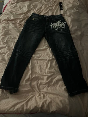 Wrung Streetlife baggy jeans med tryck - Säljer ett par mörkblå baggy jeans från Wrung Streetlife med stort vitt graffiti-tryck på ena framsidan. Jeansen har klassiska fem fickor, kontrastsömmar och coola guldfärgade detaljer vid fickorna. Perfekta för dig som gillar streetwear och loose fit.