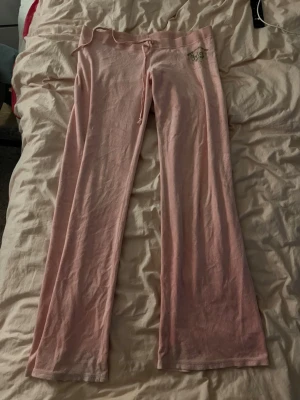 Rosa mjukisbyxor från Juicy Couture - Säljer ett par ljusrosa mjukisbyxor från Juicy Couture i storlek large. Byxorna har snörning i midjan, bootcut-snitt och ett grönt tryck på höften. Materialet är mjukt med 80% bomull och 20% polyester, perfekt för chill dagar.