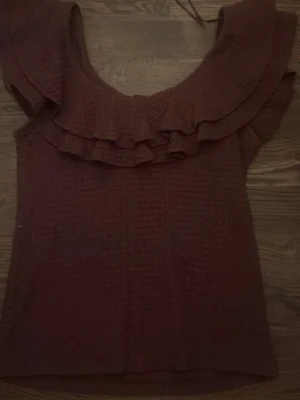 Brun volangtop från Lindex - Säljer en brun topp från Lindex i storlek M. Toppen har en snygg offshoulder-modell med dubbla volanger runt axlarna och ett strukturerat tyg som ger en cool känsla. Perfekt för dig som gillar feminina detaljer och vill ha något extra till garderoben.