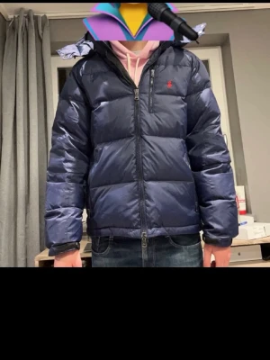 Mörkblå dunjacka från Polo Ralph Lauren - Säljer en mörkblå dunjacka från Polo Ralph Lauren med huva och dragkedja. Jackan har en klassisk puffer-look, lång ärm och ikonisk röd logga på bröstet. Perfekt för kalla dagar och riktigt snygg till jeans eller hoodie.