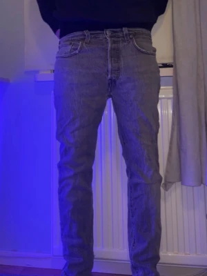 Levis 501 grå jeans straight fit - Säljer ett par klassiska Levis 501 jeans i grå tvätt. De har rak passform, fem fickor och knappgylf. Jeansen är i denim och har den ikoniska Levis-lappen bak i midjan. Perfekta för dig som gillar en tidlös och chill stil. De är i acceptabelt skick och nypriset på dessa jeans är ungefär 1000 kr. Storlek W26 L30.