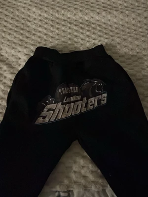 Svarta mjukisbyxor London Shooters - Säljer ett par svarta mjukisbyxor med stort tryck på framsidan där det står 'London Shooters' och ett pantermotiv. Perfekta för chill dagar eller när du vill ha något bekvämt. Resår i midjan och mjukt material.