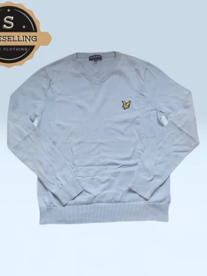 Ljusgrå stickad tröja Lyle & Scott - Snygg ljusgrå stickad tröja från Lyle & Scott med klassisk v-ringning och ikonisk gul broderad logga på bröstet. Tröjan har ribbade muddar vid ärmslut och nederkant, perfekt för dig som gillar stilrena och tidlösa plagg.