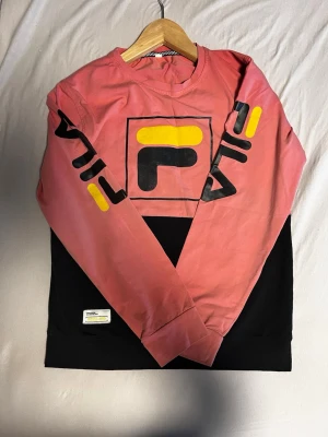 Fila långärmad tröja i rosa och svart - Säljer en långärmad tröja från Fila i rosa och svart med stor logga på bröstet och tryck på ärmarna. Tröjan har gula och svarta detaljer och är gjord i mjuk bomull. Perfekt för dig som gillar sportig och färgstark stil.