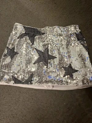 Silvrig paljettkjol med stjärnor - Kort kjol täckt av silvriga paljetter och stora svarta stjärnor. Kjolen har en elastisk midja och är fodrad med vitt tyg för extra komfort. Perfekt för dig som vill sticka ut och glänsa lite extra.