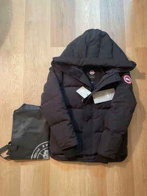 Svart pufferjacka från Canada Goose - Säljer en svart pufferjacka från Canada Goose med huva och klassisk logga på ärmen. Jackan har dragkedja och knappar framtill, samt ribbade muddar vid ärmsluten. Perfekt för kalla dagar och riktigt snygg streetstil.