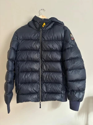 Mörkblå pufferjacka från Parajumpers - Säljer en mörkblå pufferjacka från Parajumpers, modell Sheen. Jackan har huva, ribbade muddar och dragkedja framtill. Klassisk patch på ärmen. Knappt använd. Är i st M i herrstorlek 