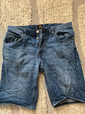 Blå jeansshorts från Lee - Säljer ett par klassiska blå jeansshorts från Lee med normal passform för män. Shortsen har fem fickor, bälteshällor och knappgylf med Lee-logga på knappen. Tillverkade i slitstark bomull med snygg tvättad look och Lee-läderpatch bak i midjan. Storlek W32