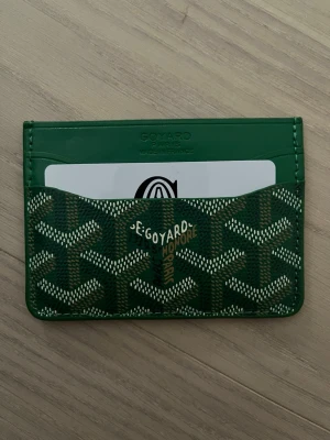 Grön korthållare  - Säljer en snygg grön korthållare deras ikoniska mönster i vitt och gult. Korthållaren har flera fack för kort och är tillverkad i skinn med ett klassiskt tryck. Perfekt för dig som vill ha något stilrent och lyxigt i fickan.